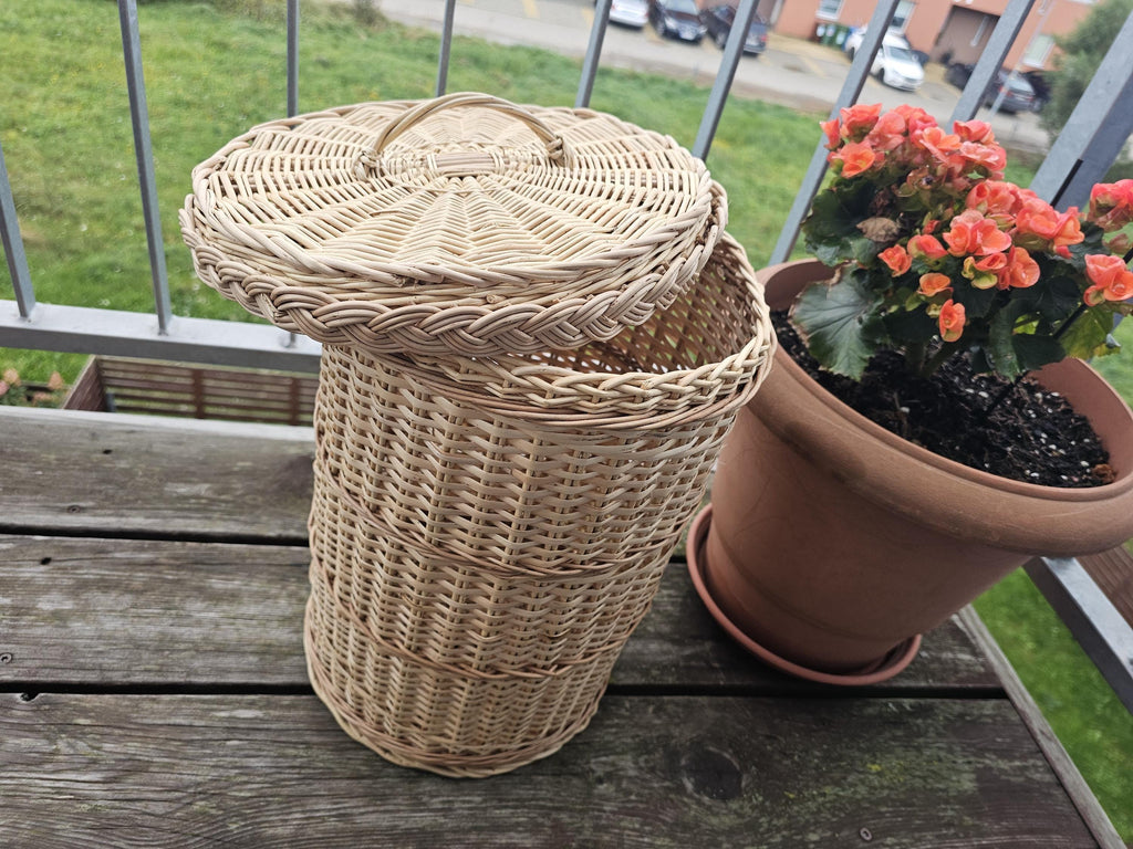 Handwoven willow trash can with lid on a wooden balcony table, lid tilted to show the plaited rim, next to a potted plant
