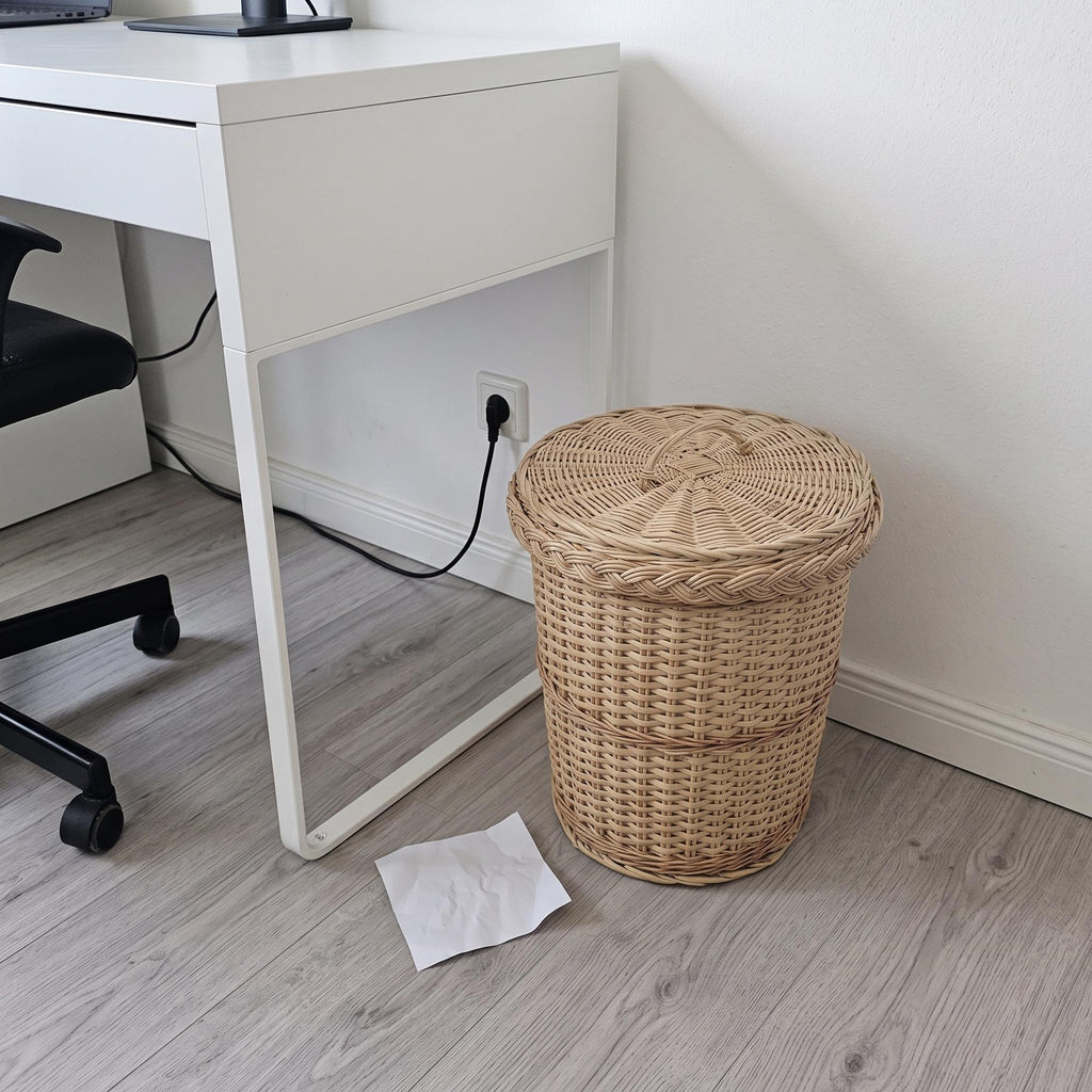 Round willow trash can with lid under a white desk in a home office, crumpled paper on the grey floor