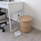 Round willow trash can with lid under a white desk in a home office, crumpled paper on the grey floor