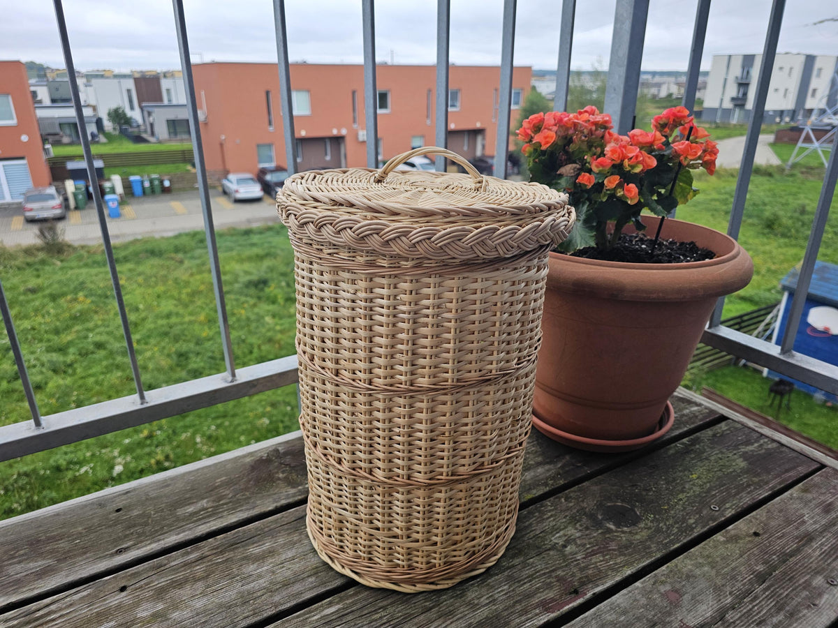 Tall round handwoven willow waste basket with lid and plaited rim, standing on a wooden balcony table beside flowers